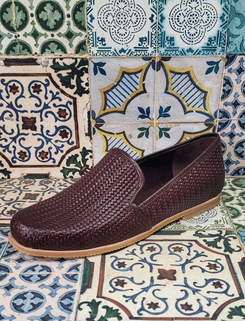 May include: A pair of brown woven slip-on shoes with a light brown sole. The shoes are on a tiled surface with a colorful pattern.