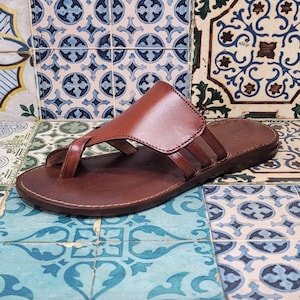 Könnte beinhalten: Braune Ledersandalen mit Zehensteg und einem einzigen Riemen über dem Fußrücken. Die Sandalen haben eine flache Sohle.