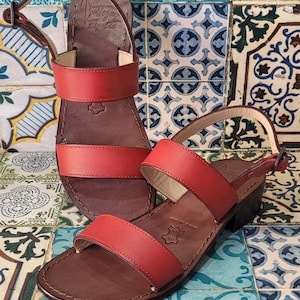 May include: Two pairs of red leather sandals with brown leather soles. The sandals have two straps across the top of the foot and an ankle strap. The sandals are on a tiled background.
