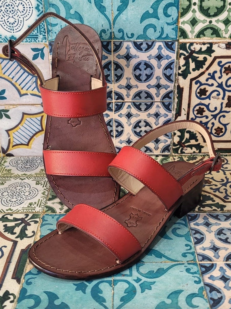 May include: Two pairs of red leather sandals with brown leather soles. The sandals have two straps across the top of the foot and an ankle strap. The sandals are on a tiled background.