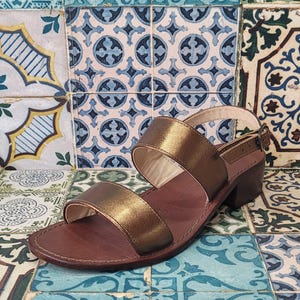 May include: Gold leather sandals with two straps and a low block heel. The sandals are on a patterned tile background.