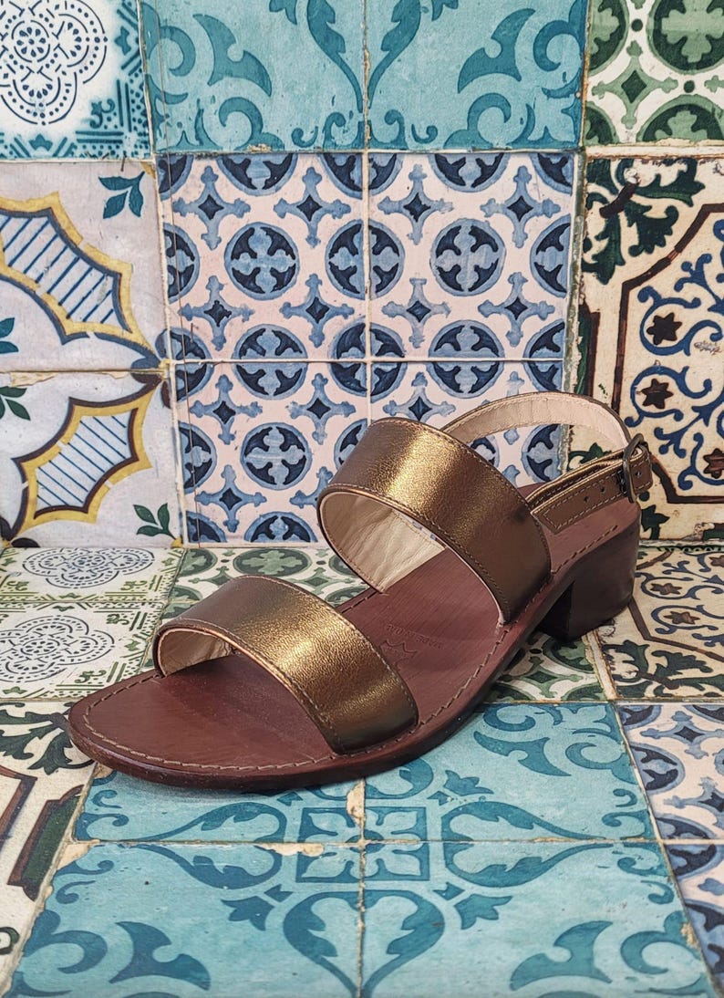 May include: Gold leather sandals with two straps and a low block heel. The sandals are on a patterned tile background.