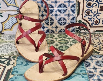 May include: Two pairs of red leather sandals with criss-cross straps and a buckle closure. The sandals are on a tiled background.