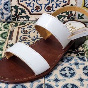 May include: White leather sandals with two straps and a brown leather sole. The sandals have a buckle closure on the ankle strap.