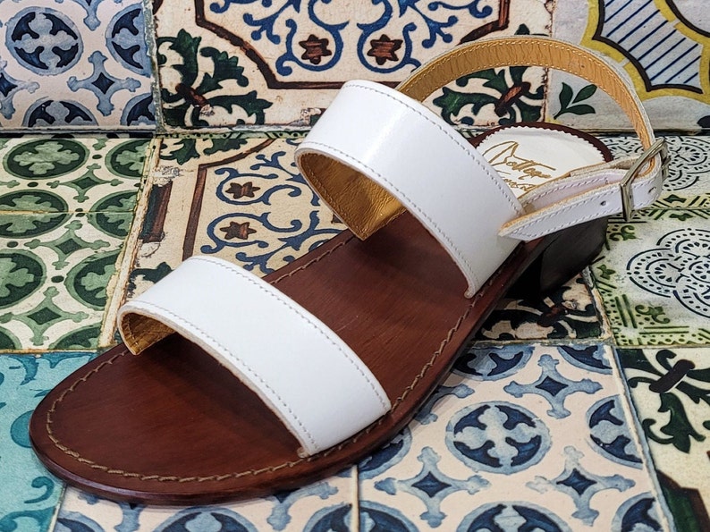 May include: White leather sandals with two straps and a brown leather sole. The sandals have a buckle closure on the ankle strap.