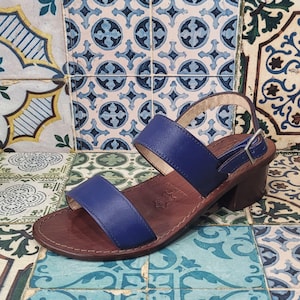 May include: Blue leather heeled sandals with two straps and a buckle closure. The sandals are on a patterned tiled floor.