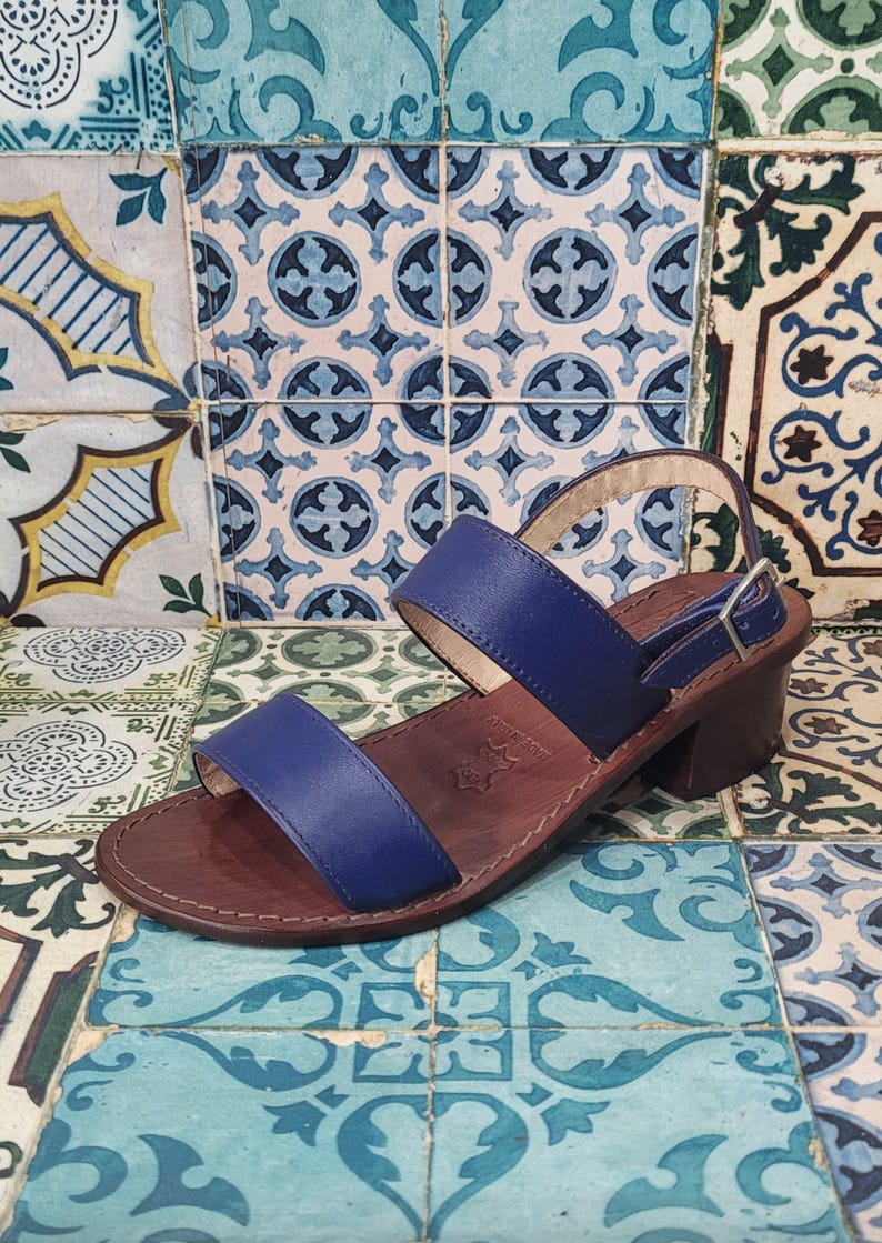 May include: Blue leather heeled sandals with two straps and a buckle closure. The sandals are on a patterned tiled floor.