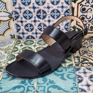 May include: Black leather sandals with two straps and a block heel. The sandals are on a patterned tiled floor.