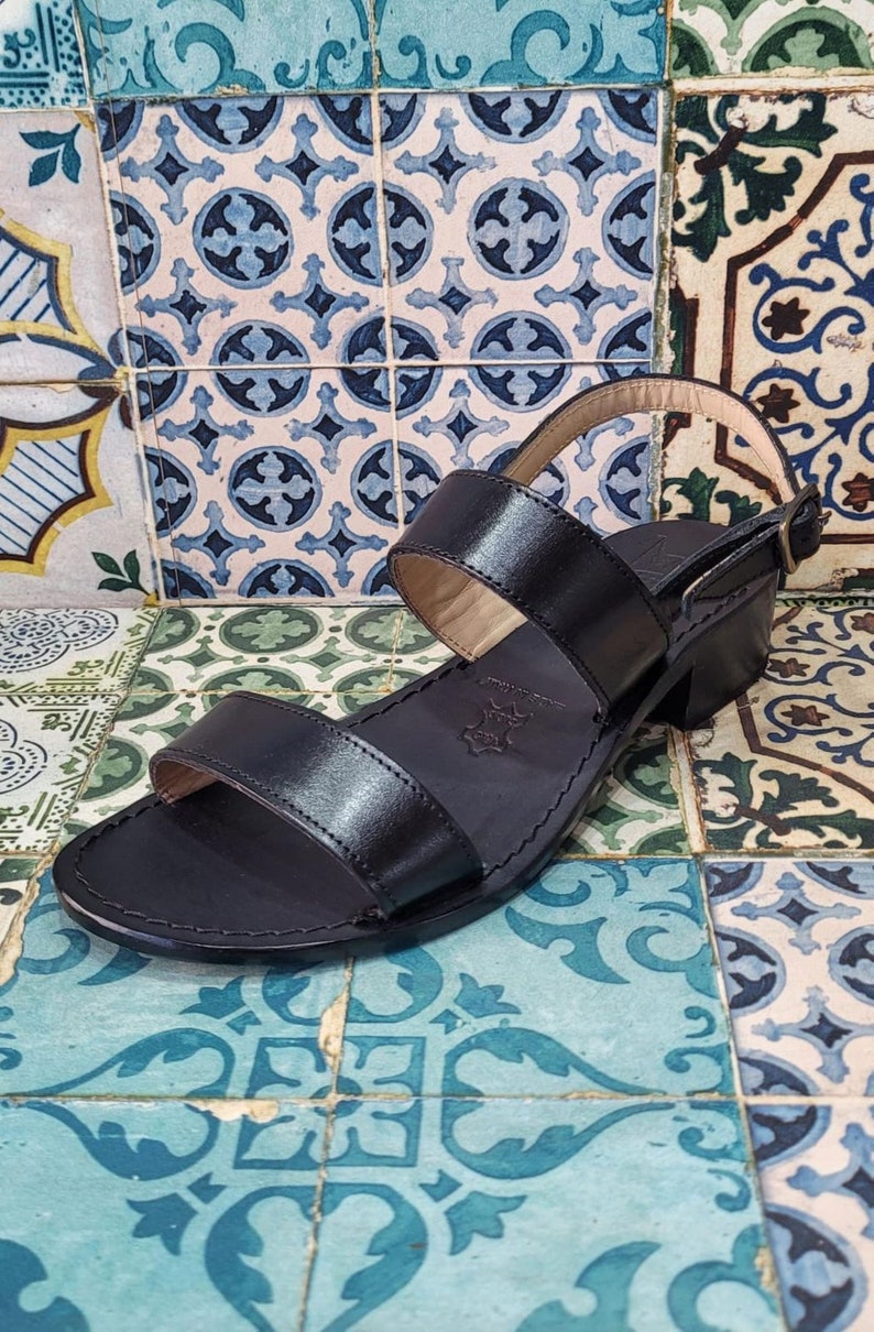 May include: Black leather sandals with two straps and a block heel. The sandals are on a patterned tiled floor.