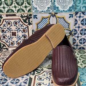 May include: A pair of brown woven leather slip-on shoes with a brown rubber sole. The shoes are on a patterned tile floor.