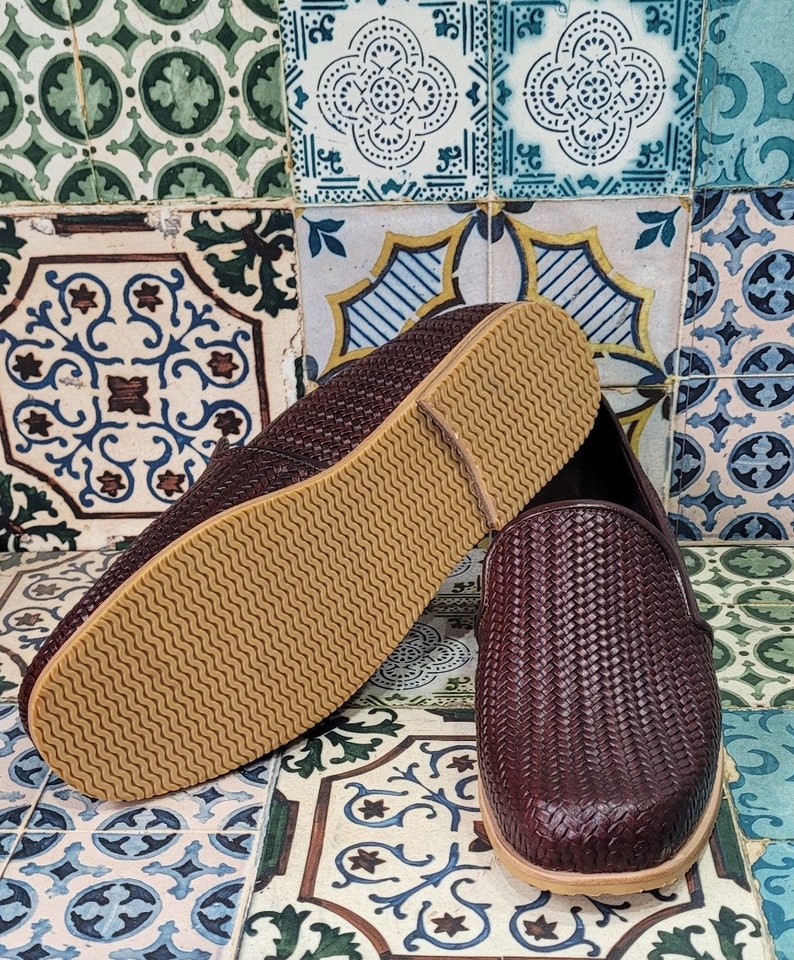 May include: A pair of brown woven leather slip-on shoes with a brown rubber sole. The shoes are on a patterned tile floor.