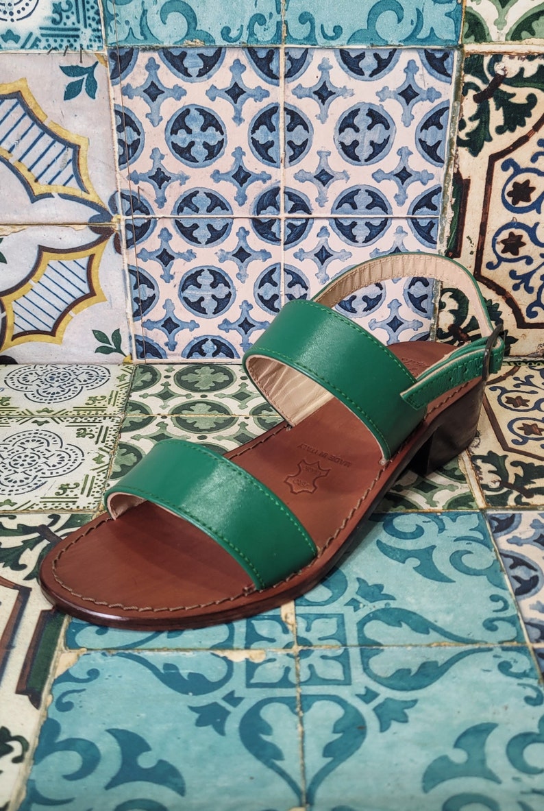 May include: Green leather heeled sandals with two straps and a brown leather sole. The sandals are on a patterned tile background.