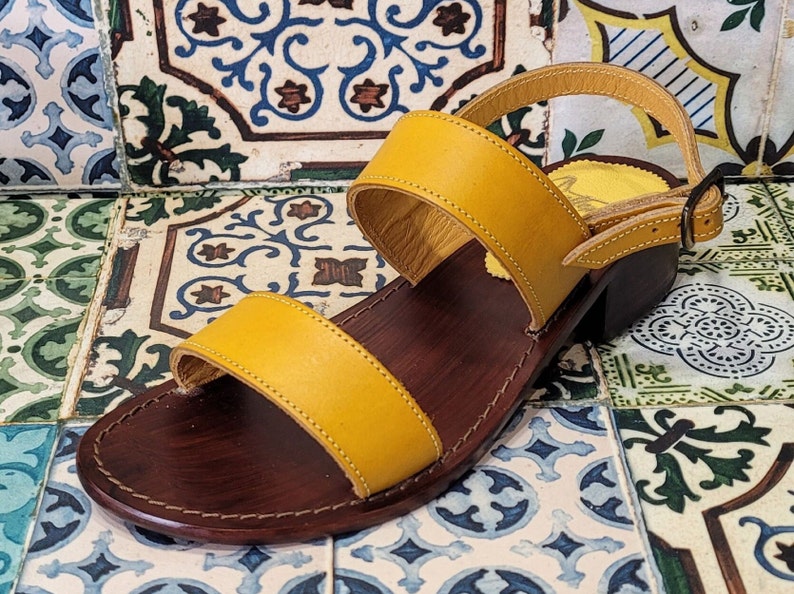 May include: A pair of yellow leather sandals with a brown leather sole and a buckle fastening. The sandals are on a tiled background.