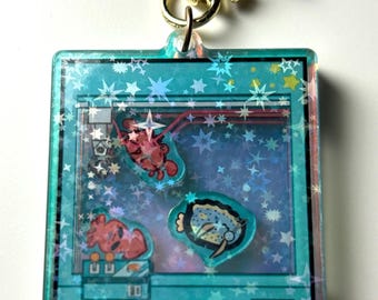 May include: A turquoise blue keychain with a gold star charm. The keychain features a clear plastic frame with a glittery background and a cartoon image of a claw machine with three prizes: a red lobster, a red crab, and a blue fish.