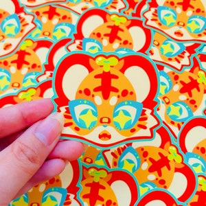 May include: A close-up of a sticker featuring a cartoon tiger with orange and red fur, wearing blue glasses with yellow stars. The tiger has a surprised expression and a yellow bow on its head.