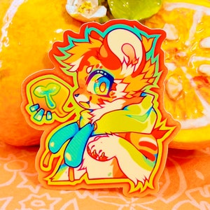 Può includere: Un adesivo colorato con un animale dei cartoni animati con pelliccia arancione, gialla e blu. L'animale tiene una foglia verde nella sua zampa. L'adesivo ha un bordo arancione brillante.