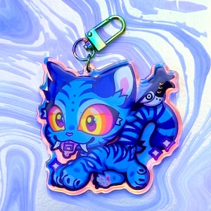 May include: A blue cat-shaped keyring with a cartoon design. The cat features rainbow eyes, sharp teeth, and a striped tail. It is attached to an iridescent metal clasp. The keyring is approximately 7 cm in length.