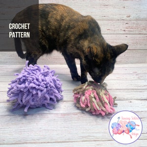 May include: Two crocheted cat toys, one purple and one pink and brown, on a wooden surface. A cat is sniffing the pink and brown toy. The text "Dancing Sisters Design" is in a circle with a yarn ball and needles.