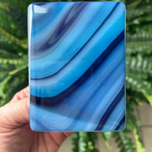 May include: A rectangular glass object with a swirling pattern of blues and white. The glass has rounded edges and a glossy finish. The design features bands of light and dark blue, creating a flowing, abstract effect. The object is held in a hand.