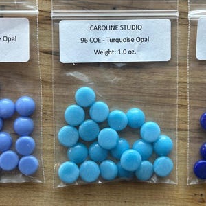 May include: Three small plastic bags filled with colorful glass beads. The bags are labeled "JCAROLINE STUDIO" with the color and weight. The beads are in shades of blue: Hydrangea Blue Opal, Turquoise Opal, and Deep Royal Blue Opal. Each bag weighs 1.0 oz.