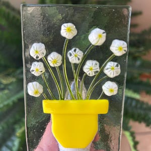May include: A glass night light featuring a yellow flower pot with white flowers and yellow centers. The flowers have green stems and are set against a clear, textured background. The night light has a white base.