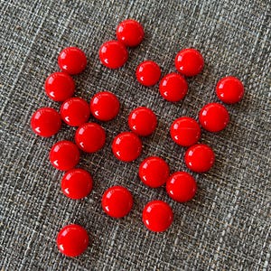 May include: A collection of twenty-one small, circular, bright red cabochons with a smooth, glossy finish. Each piece features a domed top and a flat base, arranged in a loose cluster. The objects are uniform in color and shape, reflecting light.