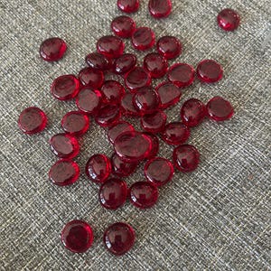May include: A collection of small, round, translucent red glass gems with a smooth, polished surface. These decorative pebbles are uniform in shape and feature a deep crimson hue that allows light to pass through, creating a vibrant appearance.