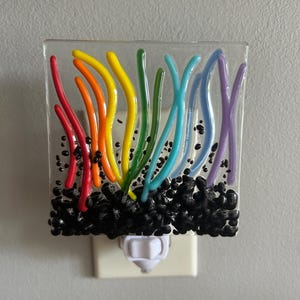 May include: A square glass night light featuring a clear base with vertical, curved, multi-colored glass lines in red, orange, yellow, green, blue, and purple. The bottom section contains a dense cluster of small, black, rounded glass beads.
