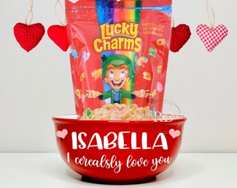 May include: Red ceramic bowl with the words "ISABELLA I cerealsly love you" and two pink hearts. A box of Lucky Charms cereal is sitting on top of the bowl.