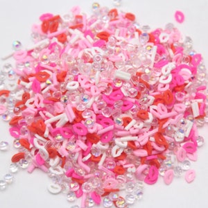 May include: A mix of pink, red and white sprinkles in the shape of lips, hearts and lines. The sprinkles are mixed with clear iridescent beads.