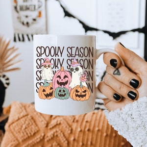 May include: White ceramic mug featuring the text "Spooky Season" stacked vertically. The mug is decorated with cartoon ghosts and pumpkins wearing witch hats and sunglasses. The mug is held by a person.