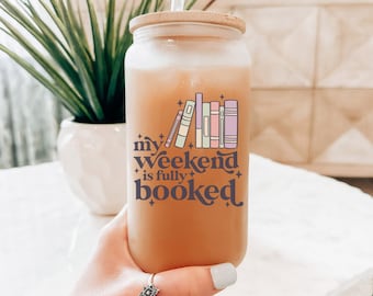 May include: A clear glass tumbler with a bamboo lid and a clear straw, filled with a light brown beverage. The tumbler features the text "my weekend is fully booked" with a graphic of colorful books. The tumbler is held by a person.