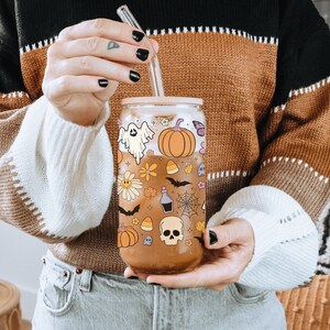 Pu&ograve; includere: Un bicchiere di vetro trasparente con coperchio in bamb&ugrave; e cannuccia in vetro, decorato con illustrazioni a tema Halloween. Il design include un fantasma, una zucca, fiori, pipistrelli e un teschio. Il bicchiere &egrave; riempito con un liquido marrone.