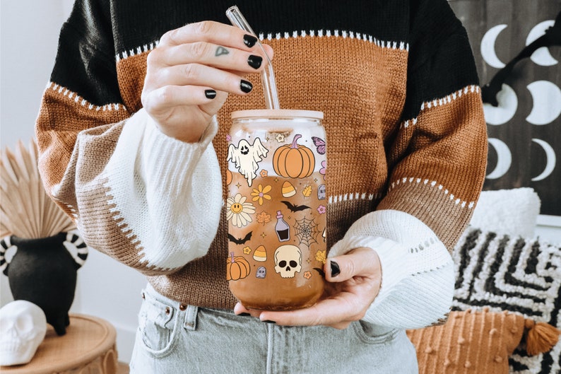 Pu&ograve; includere: Un bicchiere di vetro trasparente con coperchio in bamb&ugrave; e cannuccia in vetro, decorato con illustrazioni a tema Halloween. Il design include un fantasma, una zucca, fiori, pipistrelli e un teschio. Il bicchiere &egrave; riempito con un liquido marrone.