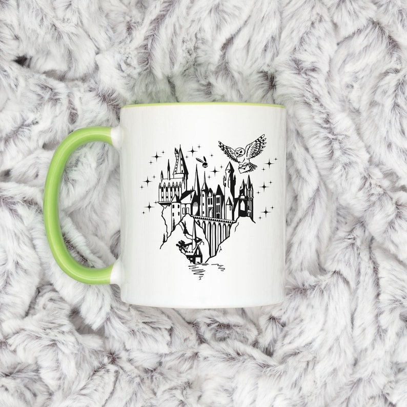 Puede incluir: Taza de cer&aacute;mica blanca con asa y borde verdes. La taza presenta una ilustraci&oacute;n negra de un castillo, un b&uacute;ho que lleva una carta y estrellas. El dise&ntilde;o recuerda un tema de fantas&iacute;a.