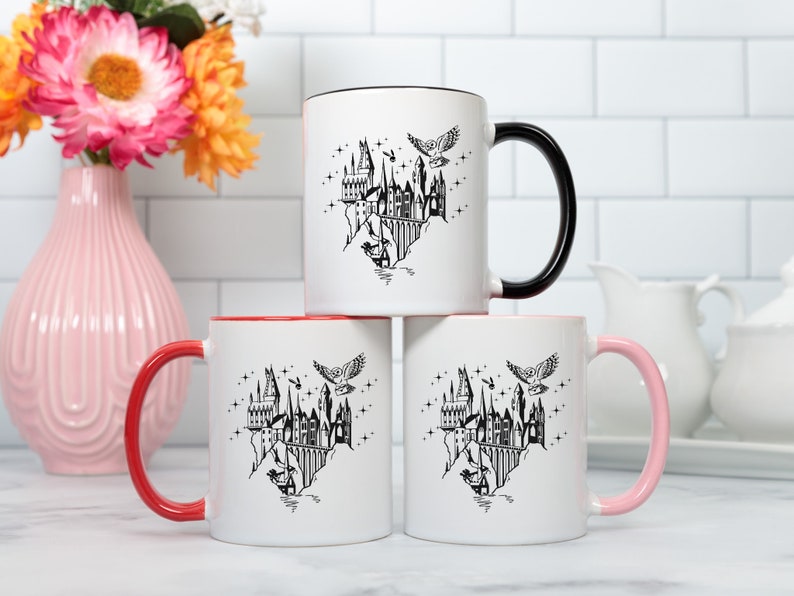 Puede incluir: Tres tazas de cer&aacute;mica blancas con detalles en negro, rojo y rosa. Cada taza presenta una ilustraci&oacute;n negra de un castillo, un b&uacute;ho y estrellas. Las tazas est&aacute;n apiladas, con un jarr&oacute;n de flores en el fondo.