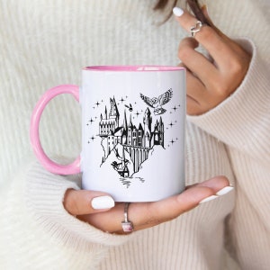 Puede incluir: Taza de cer&aacute;mica blanca con asa e interior rosa. La taza presenta una ilustraci&oacute;n negra de un castillo, un b&uacute;ho que lleva una carta y estrellas. La taza est&aacute; sostenida en las manos de una persona.