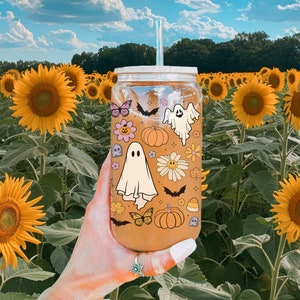 Pu&ograve; includere: Un bicchiere di vetro trasparente con cannuccia, decorato con adesivi a tema Halloween, tra cui fantasmi, pipistrelli, zucche e fiori. Il bicchiere &egrave; tenuto davanti a un campo di girasoli sotto un cielo blu.