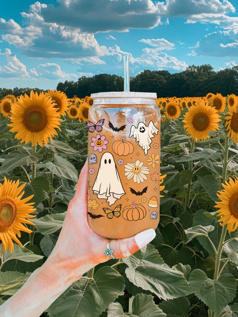 Pu&ograve; includere: Un bicchiere di vetro trasparente con cannuccia, decorato con adesivi a tema Halloween, tra cui fantasmi, pipistrelli, zucche e fiori. Il bicchiere &egrave; tenuto davanti a un campo di girasoli sotto un cielo blu.