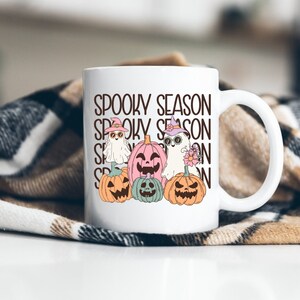 May include: White ceramic mug featuring the text "Spooky Season" and illustrations of ghosts and pumpkins. The ghosts wear hats and sunglasses, and the pumpkins are in shades of orange, pink, and green. The mug has a white handle.