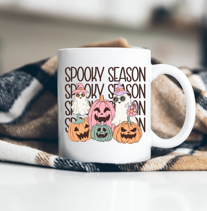 May include: White ceramic mug featuring the text "Spooky Season" and illustrations of ghosts and pumpkins. The ghosts wear hats and sunglasses, and the pumpkins are in shades of orange, pink, and green. The mug has a white handle.