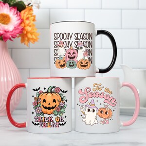 May include: Three white ceramic mugs with Halloween-themed designs. One mug has a black handle and the text "Spooky Season" with cartoon ghosts and pumpkins. Another mug has a red handle with a pumpkin and the text "Trick or Treat". The third mug has a pink handle with a ghost and pumpkin and the text "'Tis the Season".