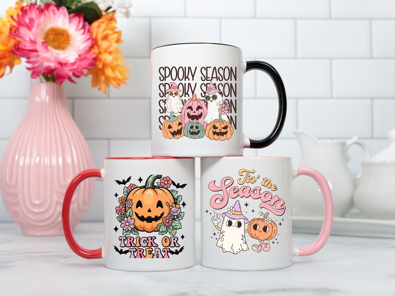 May include: Three white ceramic mugs with Halloween-themed designs. One mug has a black handle and the text "Spooky Season" with cartoon ghosts and pumpkins. Another mug has a red handle with a pumpkin and the text "Trick or Treat". The third mug has a pink handle with a ghost and pumpkin and the text "'Tis the Season".