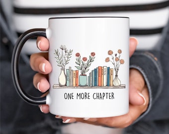 May include: White ceramic mug with a black handle and rim, featuring a bookshelf design with vases of flowers and the text "ONE MORE CHAPTER". The mug is held by a person.