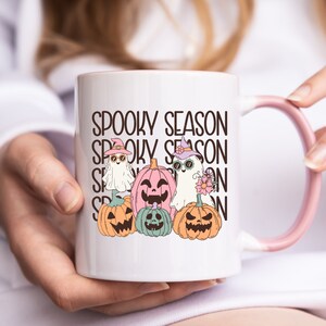 May include: White ceramic mug with a pink handle and rim, featuring the text "Spooky Season" stacked vertically. The mug is decorated with cartoon ghosts and jack-o'-lanterns in pastel colors, perfect for Halloween.