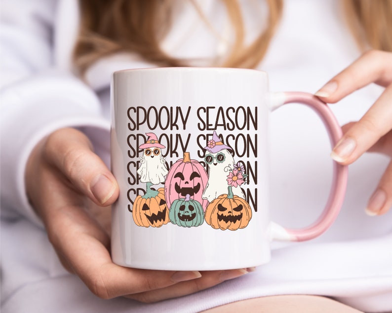 May include: White ceramic mug with a pink handle and rim, featuring the text "Spooky Season" stacked vertically. The mug is decorated with cartoon ghosts and jack-o'-lanterns in pastel colors, perfect for Halloween.