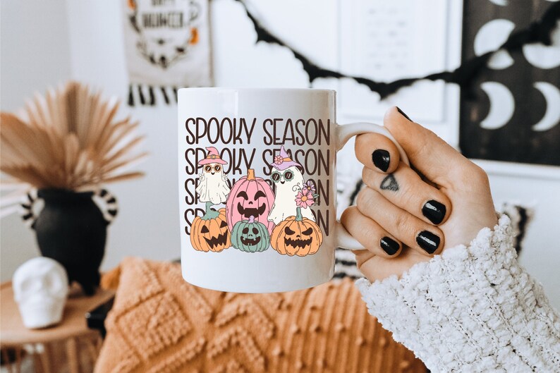 May include: White ceramic mug featuring the text "Spooky Season" stacked vertically. The mug is decorated with cartoon ghosts and pumpkins wearing witch hats and sunglasses. The mug is held by a person.