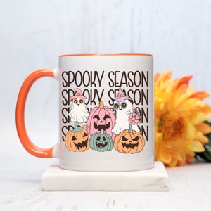 May include: White ceramic mug with an orange handle and rim, featuring a "Spooky Season" design. The design includes cartoon ghosts and jack-o'-lanterns in pastel colors. A perfect gift for Halloween lovers.