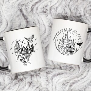 Puede incluir: Dos tazas de cer&aacute;mica blanca con asas y bordes negros. Una taza presenta una ilustraci&oacute;n negra del castillo de Hogwarts con un b&uacute;ho y estrellas. La otra taza tiene un dise&ntilde;o circular con el texto "Hogwarts is my home" y una ilustraci&oacute;n del castillo.