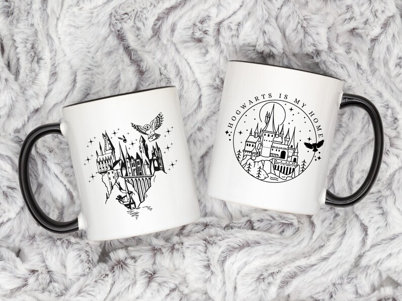 Puede incluir: Dos tazas de cer&aacute;mica blanca con asas y bordes negros. Una taza presenta una ilustraci&oacute;n negra del castillo de Hogwarts con un b&uacute;ho y estrellas. La otra taza tiene un dise&ntilde;o circular con el texto "Hogwarts is my home" y una ilustraci&oacute;n del castillo.